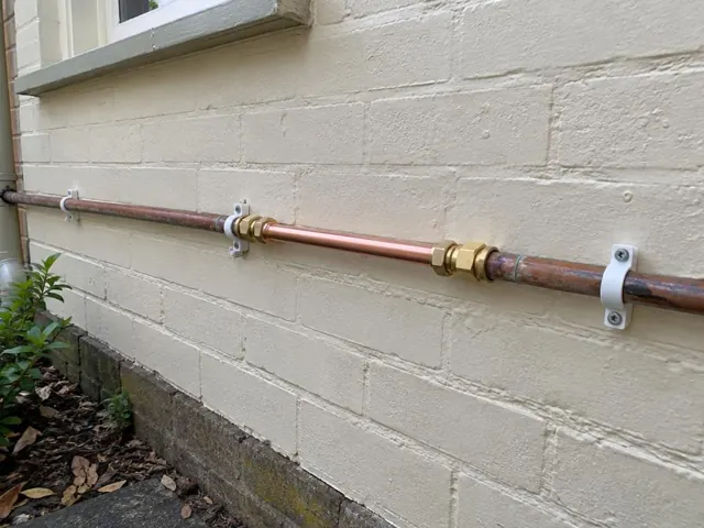 Repaired copper water pipe mounted along a painted brick exterior wall in Lindfield